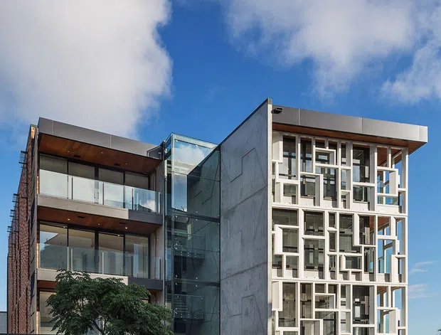 Street-facing photograph of a contemporary multi-storey apartment or mixed-use building. The facade combines exposed concrete, extensive glazing, dark metal framing, warm timber soffits and balconies, and a distinctive white geometric screen across the right-hand elevation. A glazed vertical circulation core links the two main volumes, with a low stone-filled wall at street level. The building sits in an urban streetscape under a partly cloudy blue sky.