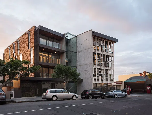 Street-level exterior photograph of a contemporary multi-storey urban building, combining exposed red brick, dark metal-framed glazing, concrete panels, and a prominent glass stair or circulation tower. The facade includes a decorative grid of projecting window frames and screens. Adjacent low-rise buildings, parked cars, and a paved street are visible, with warm evening light and a partly cloudy sky.