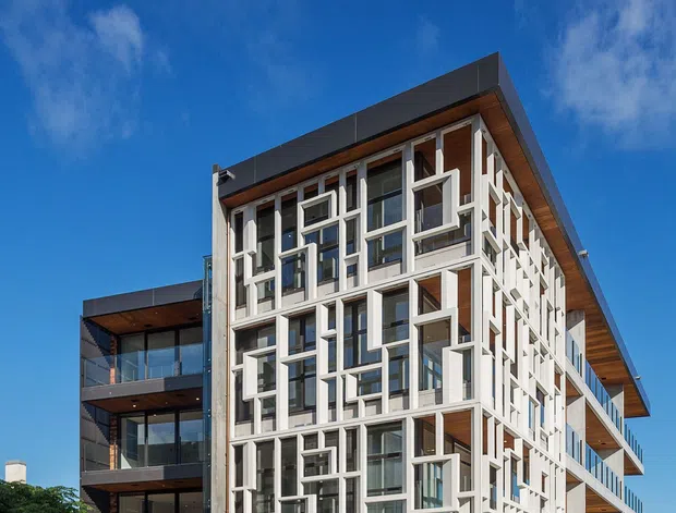 Exterior architectural photograph of a contemporary multi-storey apartment building in an urban setting. The facade features extensive glazing, projecting white geometric window surrounds, dark metal cladding, warm timber soffits, balconies with glass balustrades, and a textured stone retaining base. A street and partial waterfront or open horizon are visible in the background.