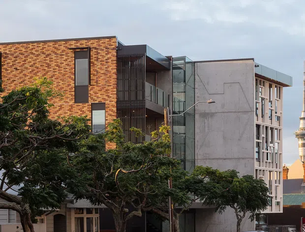 Urban exterior view of a contemporary multi-storey building complex, combining a warm patterned brick facade with exposed concrete, dark metal screening, recessed balconies and extensive glazing. Mature street trees partially obscure the lower levels. A tall landmark tower is visible in the background, reinforcing the dense city setting.