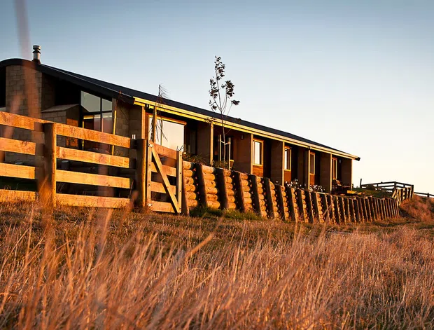 A low, elongated rural house viewed from the exterior at sunset, set among dry grasses behind a timber post-and-rail fence. The home has dark corrugated-looking pitched roof forms, warm timber cladding and exposed timber verandah elements, with a brick chimney and a long row of windows and doors opening onto a deck. The image has a warm, rustic architectural character and appears to be a single-storey dwelling.