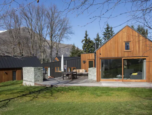 Exterior view of a compact cluster of timber-clad buildings set in a wooded mountain landscape. The main gable-roofed structure has warm vertical timber siding, a large glazed opening, and a small upper window. A dark metal-roofed connecting volume, covered timber deck, outdoor dining furniture, and a substantial stone fireplace or chimney are visible.