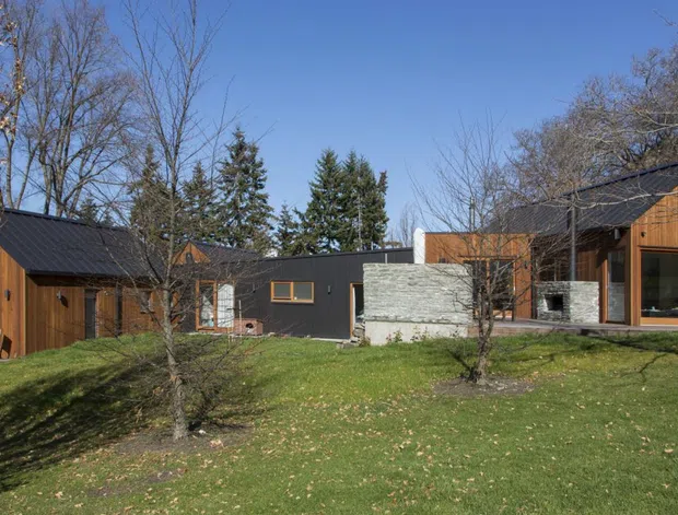 A low-rise contemporary residential compound set within a landscaped, wooded site. Several connected or closely spaced buildings feature dark pitched metal roofs, warm vertical timber cladding, dark painted wall sections, exposed concrete masonry, and large timber-framed windows and doors. The foreground is a broad lawn with mature deciduous trees and evergreen plantings under a clear blue sky.
