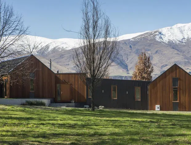 Single-storey contemporary residential building composed of several dark timber-clad gabled volumes, set in a broad lawn with leafless trees and dramatic snow-covered mountains in the background. The low, elongated form and simple vertical cladding create a restrained rural alpine composition.