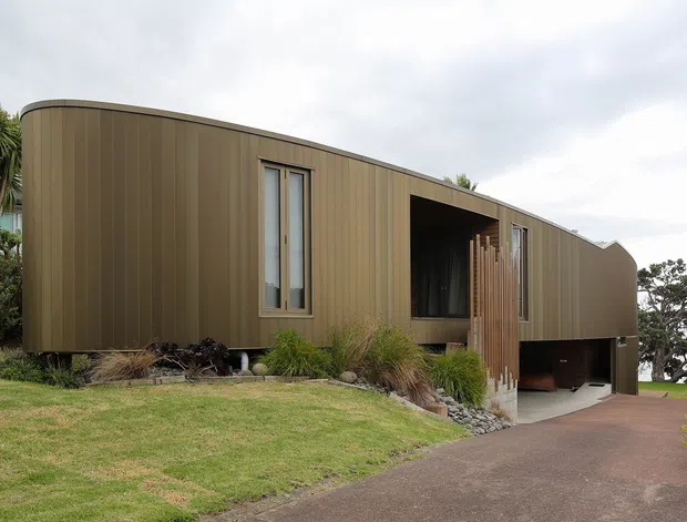 A contemporary curved single-storey house clad in vertically ribbed warm brown panels, set on a landscaped sloping site. The facade features narrow windows, a recessed entry porch with vertical timber screening, and a partially open undercroft or garage area. A paved driveway approaches the building, with lawn and ornamental planting around it.
