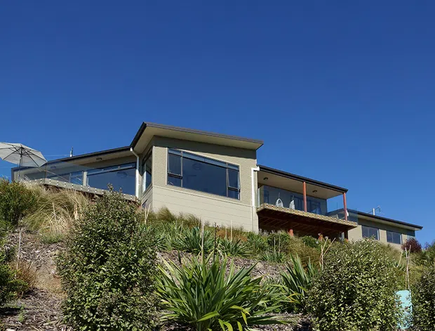 Exterior view of a modern multi-level house set into a landscaped sloping site. The home has pale horizontal cladding, broad dark-framed windows, projecting roof planes, and a timber balcony or deck, with native-style planting on the hillside.