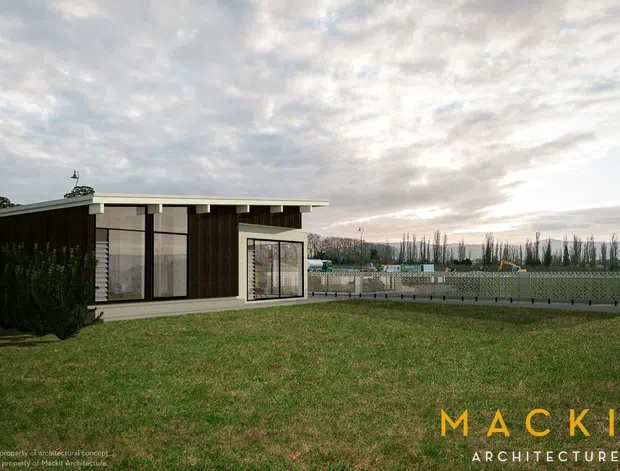 Exterior view of a low-profile modern single-storey building set in an open grassy landscape. The structure features dark vertical cladding, extensive floor-to-ceiling glazing, a shallow flat roof with broad overhangs, and a pale perforated or patterned boundary wall enclosing a terrace or pool-like area. The image has a restrained contemporary architectural character under an overcast sky.