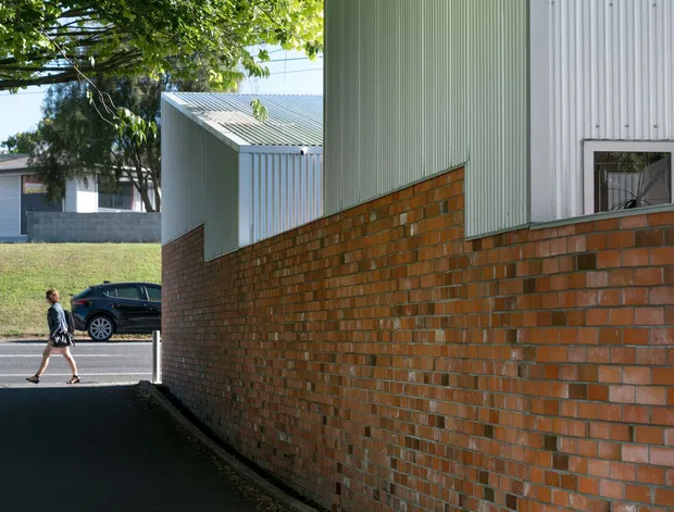 Exterior architectural view of a narrow paved passage beside a low brick wall and a tall building clad in vertically ribbed pale green and white metal panels. A smaller matching structure, street frontage, mature trees, and a pedestrian are visible beyond. The image emphasizes the building envelope, brickwork, and corrugated metal cladding in a leafy urban setting.