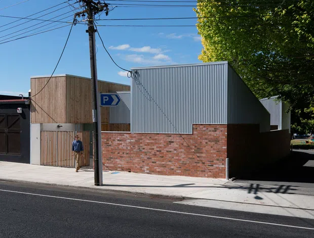 Street-level exterior view of a compact contemporary commercial or institutional building. The facade combines a low red brick wall, vertically ribbed grey metal cladding, and a timber-slatted entrance section. A parking sign, utility pole, overhead wires, paved footpath, roadway, and mature street tree are visible. Bright clear daylight emphasizes the crisp geometric massing and material contrast.