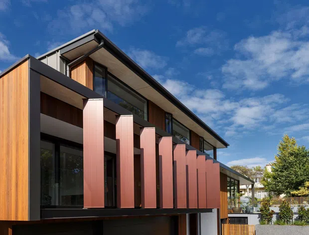Exterior architectural photograph of a contemporary multi-level residence with strong horizontal rooflines, dark framing, warm timber cladding, and a rhythmic row of projecting red-brown vertical fins forming a balcony or facade screen. Large glazed openings and a lower enclosed garage or undercroft are visible. The home sits in a landscaped suburban setting beneath a blue sky.