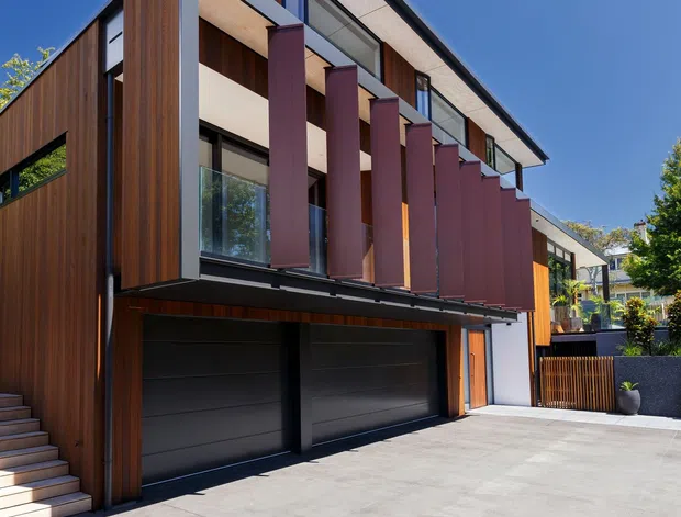 A contemporary multi-storey residential facade viewed from the driveway. The home features warm vertical timber cladding, dark framed glazing, projecting upper-level balconies with burgundy privacy screens, a sharply pitched roof, and an integrated double garage. A broad concrete driveway and timber privacy gate occupy the foreground.