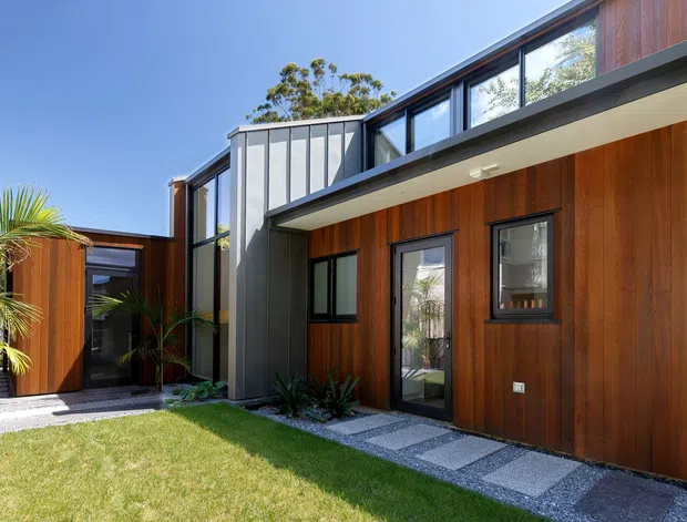 Exterior view of a contemporary multi-level residence arranged around a landscaped courtyard. The facade combines warm vertical timber cladding with dark metal-framed glazing and a projecting upper volume, while a gravel-and-concrete stepping-stone path crosses the lawn toward the entry. Tropical planting and a clear blue sky contribute to the bright outdoor setting.