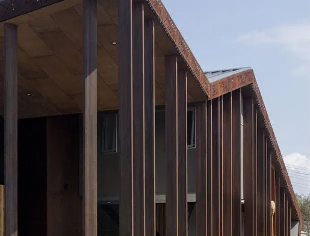 Exterior view of a long, narrow contemporary building with a dramatic sloping roof and deep covered veranda. Repeated dark timber or metal-clad vertical columns create a strong rhythmic facade, while decorative patterned fascia panels run along the roof edge and base. The structure appears raised slightly above a gravel and lawn landscape, with glazed openings and a recessed entry visible between the columns.
