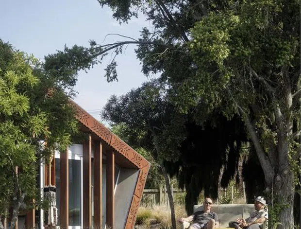 A contemporary pavilion-like building set within a leafy landscaped garden, featuring a sharply angled terracotta-toned exterior frame with decorative geometric detailing, extensive floor-to-ceiling glazing, and vertical timber elements. Two people sit on a built-in outdoor bench beneath mature trees. Stone retaining walls and natural planting surround the structure.