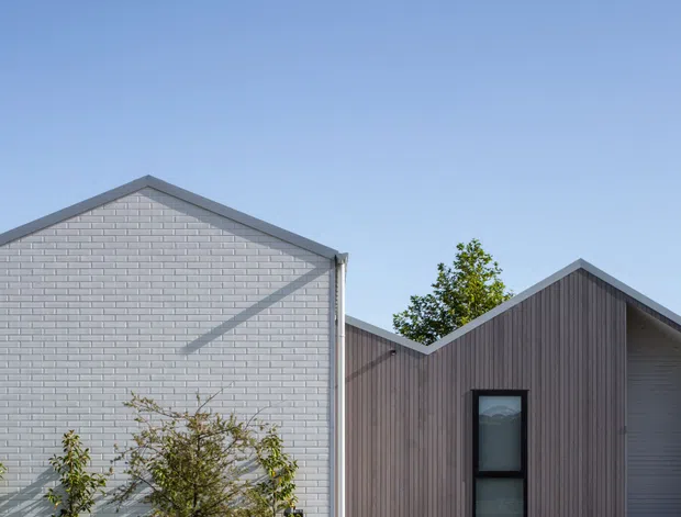 Exterior architectural view of a contemporary single-storey building composed of intersecting gabled volumes. The facade combines pale painted brick with vertical dark timber-like cladding, dark-framed windows, and simple grey roof edging. Clear blue sky and some planting surround the structure.