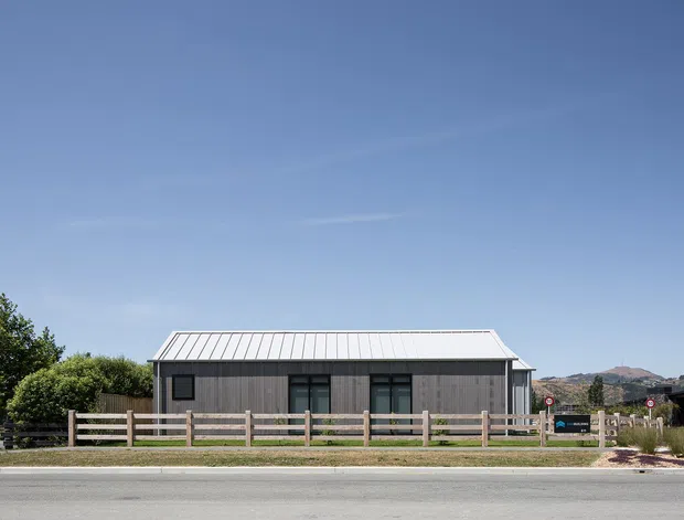 A single-storey contemporary building viewed from across a road, with a simple rectangular form, dark vertical timber-style cladding, a light standing-seam metal gable roof, and dark-framed windows and doors. A low timber post-and-rail fence runs along the frontage, with landscaped planting and distant hills behind the property.