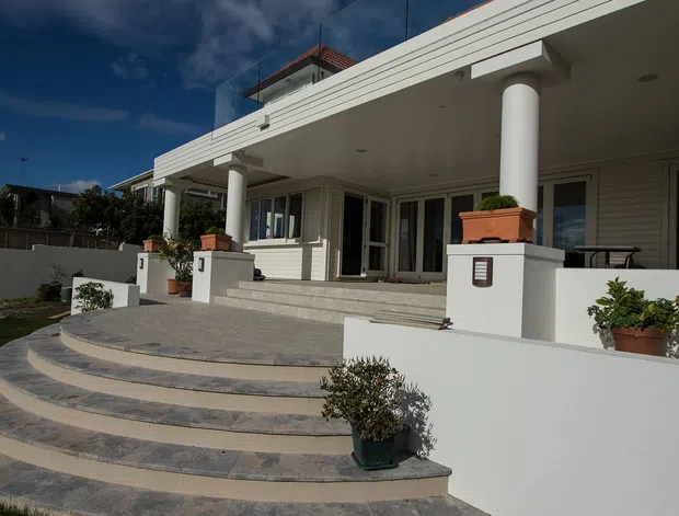 Exterior view of a light-coloured, single-storey residence with a broad covered veranda supported by round columns, glazed doors and windows, raised planter boxes, and a sweeping curved set of tiled steps leading to the lawn.