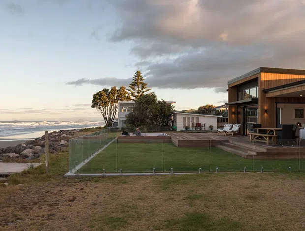 Coastal residential property viewed from the beach, featuring a contemporary dark-clad house with warm timber soffits and detailing, extensive glazing, a low flat roofline, and a fenced lawn extending toward a rocky shoreline. The image is captured at dusk with warm interior and exterior lighting.