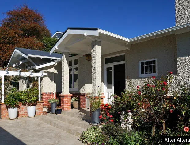 Exterior view of a single-storey suburban home with a low-pitched, multi-gabled roof and textured light-grey rendered walls. The entrance porch features square columns with brick bases, white-framed glazing and doors, and brick planter walls. Paved steps and a paved forecourt lead to a landscaped garden with rose bushes and potted plants.
