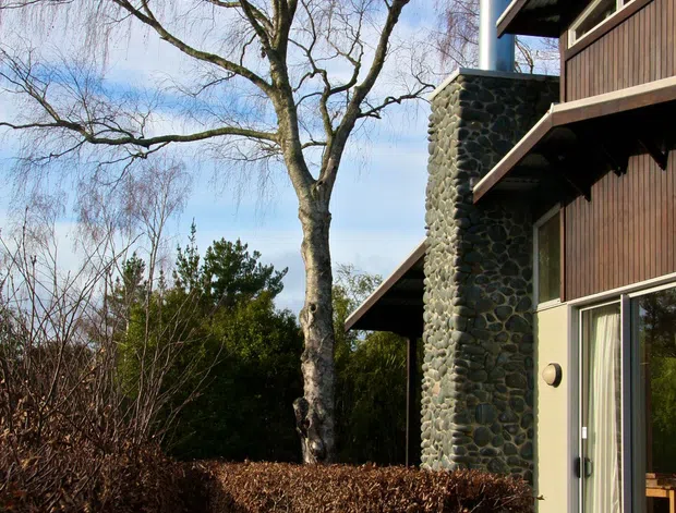 Exterior view of a modern timber-clad house set among mature trees and hedges. The building features dark vertical timber wall cladding, broad roof overhangs, large glazed doors and windows, and a prominent rounded dark-stone chimney with a metal flue. A timber deck and dense landscaped hedging occupy the foreground.