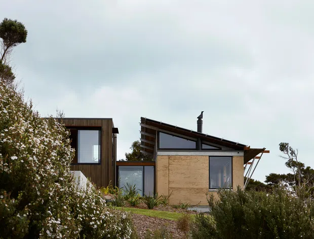 Exterior view of a low-rise contemporary dwelling set within native coastal-looking vegetation. The composition shows a dark timber-clad cubic volume at left connected to a central glazed link and a larger earthen-toned masonry or rendered volume at right. The right-hand section has a sloping metal roof with exposed eaves, a chimney, and large dark-framed windows. Wild planting and gravel landscaping surround the house beneath an overcast sky.