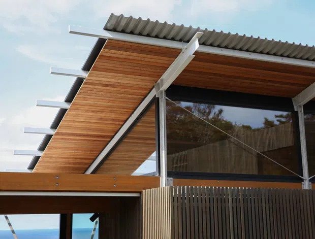 Exterior architectural detail of a contemporary building with a dramatic sloping timber-lined soffit, exposed white structural beams, corrugated roof sheeting, dark-framed glazing, and vertical timber screening. The elevated structure and ocean-like blue view visible beneath suggest a coastal setting, though the main focus is the building envelope.