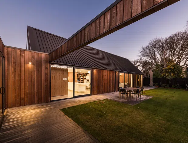 Twilight exterior view of a contemporary timber-clad residence arranged around a landscaped lawn and timber deck. The building features steep dark metal pitched roofs, a prominent elevated timber-clad link or bridge, large glazed openings, and warm interior lighting visible through the doors and windows. An outdoor dining setting sits on the deck beside the lawn.