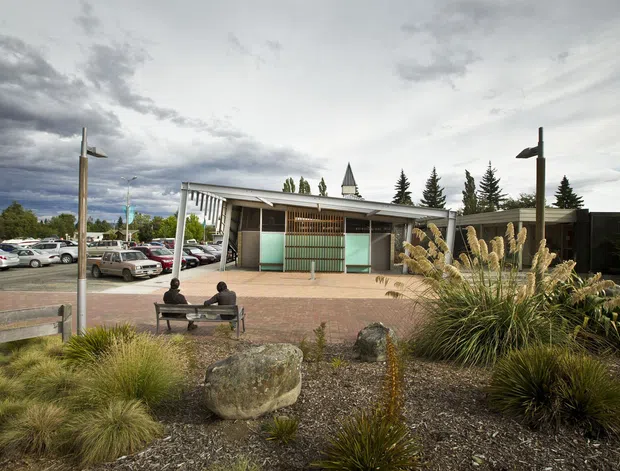 Wide exterior view of a low-rise contemporary building and landscaped forecourt. The building has a shallow roof, white structural framing, large glazed openings, timber screening, and a small projecting tower or steeple-like roof element. A paved pedestrian area with benches and modern light poles sits in front, bordered by ornamental grasses, rocks, and gravel. A car park is visible to the left under an overcast sky.
