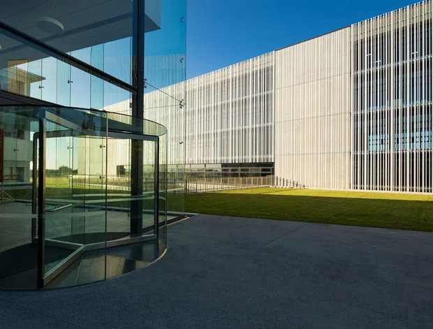 Exterior view of a contemporary institutional or commercial building arranged around a landscaped courtyard. A curved glass revolving entrance and extensive floor-to-ceiling glazing sit in the foreground, while the main facade features dense vertical metal screening over a light-coloured wall. Broad concrete paving and a neatly maintained lawn create a formal, minimalist setting under a clear blue sky.