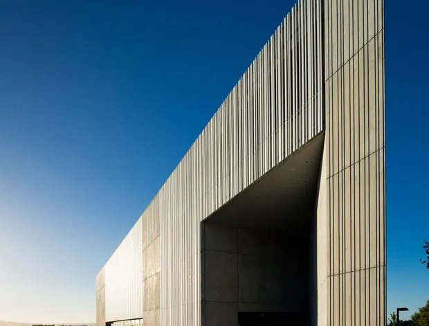 Exterior architectural photograph of a large, angular institutional or commercial building. The facade is clad in pale concrete or precast panels with strong vertical ribbing, forming a tall wedge-like volume and a dramatic recessed vehicle or entry opening. A broad paved forecourt and minimal fencing surround the structure under a clear deep-blue sky.