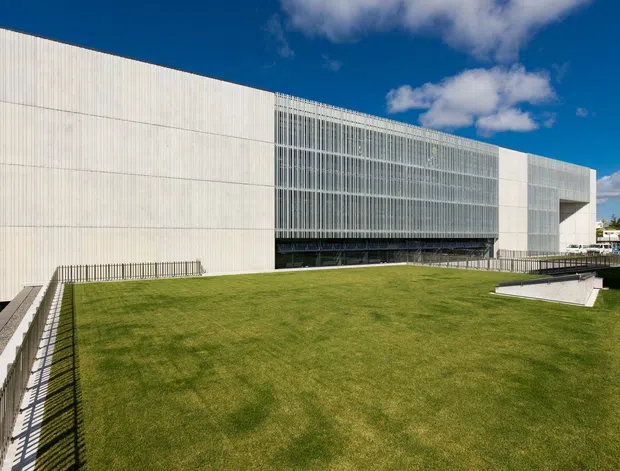 Exterior view of a large contemporary institutional or commercial building surrounding a broad elevated lawn. The facade combines pale vertical ribbed wall panels with a prominent multi-storey glazed curtain wall and projecting rectangular volumes. Walkways with metal railings frame the foreground, beneath a bright blue sky.