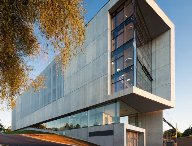 Exterior architectural photograph of a large contemporary multi-storey building on a sloped urban site. The building features a crisp rectilinear form, pale grey vertically textured cladding, extensive floor-to-ceiling glazing, a recessed glazed circulation or atrium void, and a dramatic projecting upper volume. Dark asphalt paving and a loading or service area occupy the foreground, with mature trees beside the building.