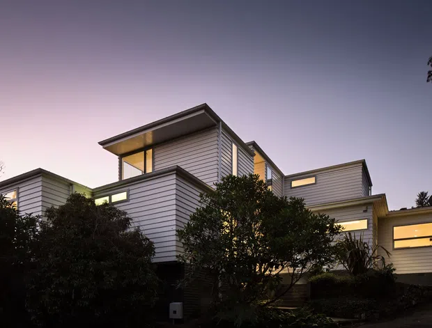Twilight exterior view of a contemporary multi-level residence composed of several articulated box-like volumes. The building features horizontal light-coloured weatherboard cladding, dark metal roof edges and window frames, and warm illumination through broad rectangular windows. Dense garden planting partially screens the lower facade.