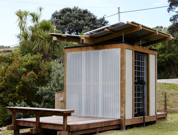 Small standalone timber-clad garden structure with a raised timber deck, translucent corrugated wall panels, a narrow glazed door or window, and a distinctive projecting roof with exposed timber framing and corrugated roofing. It sits in a landscaped grassy setting with mature planting and fencing beyond.