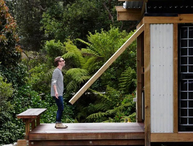 Exterior view of a compact timber-clad structure set within dense lush vegetation. A raised timber deck with a simple bench leads to the building, which features exposed timber framing, corrugated metal roofing, a large dark-framed glazed opening, and a projecting timber element. A person stands on the deck for scale.