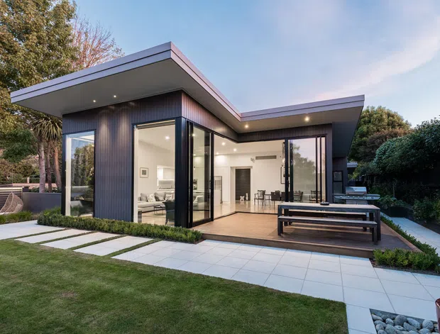 Contemporary single-storey pavilion-style home with a dramatic flat roof, dark vertical cladding, extensive floor-to-ceiling glazing, and an open-plan interior visible through the glass. The house opens onto a paved terrace with built-in bench seating, surrounded by manicured lawn, planting, and a landscaped garden.