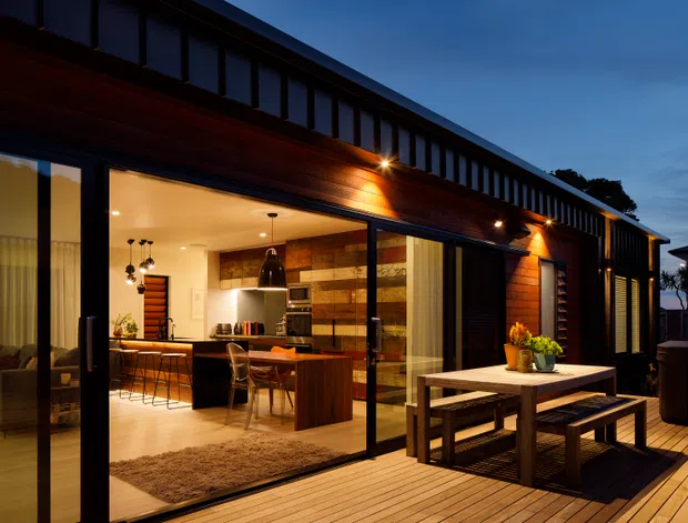 Twilight exterior view of a contemporary single-storey home extension with dark vertical cladding, warm timber soffit and decking, and extensive floor-to-ceiling glazed sliding doors. The open glazing reveals a warm kitchen and living area, while a timber outdoor dining table with benches sits on the deck.