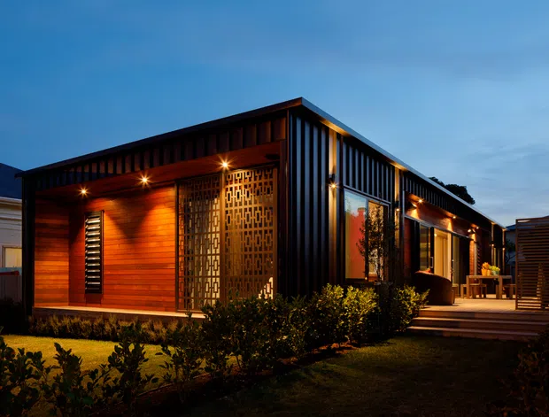 Twilight exterior view of a compact modern single-storey dwelling with a low-pitched roof, dark vertical metal cladding and warm timber wall panels. A patterned privacy screen, recessed outdoor lighting, covered entry/terrace, steps and landscaped garden are visible.