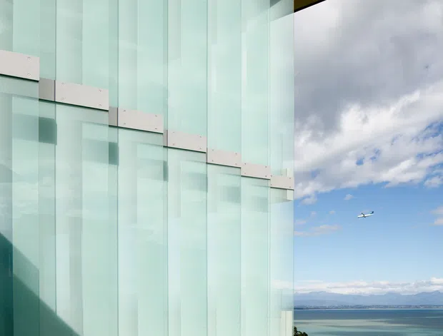 Close-up architectural exterior view of a contemporary building facade composed of tall translucent pale aqua glass panels with regularly spaced vertical divisions and light metal fixing bands. The glazing occupies the left side of the image, while a bright coastal or waterfront landscape with blue water, distant mountains, and sky is visible to the right. The composition emphasizes the building envelope and reflective/translucent facade.