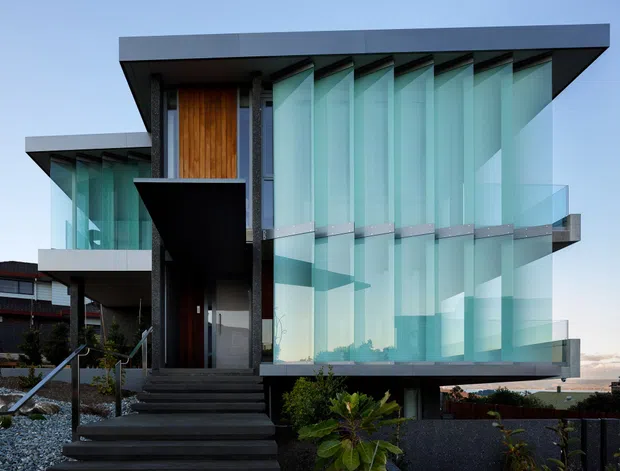 Exterior view of a striking contemporary multi-storey residence with extensive turquoise-tinted floor-to-ceiling glazing, projecting dark flat roof planes, glass balustrades, dark cladding, and a warm timber-clad entry feature. A stepped path leads to the recessed front entrance, with landscaped planting and neighbouring buildings visible around the property.