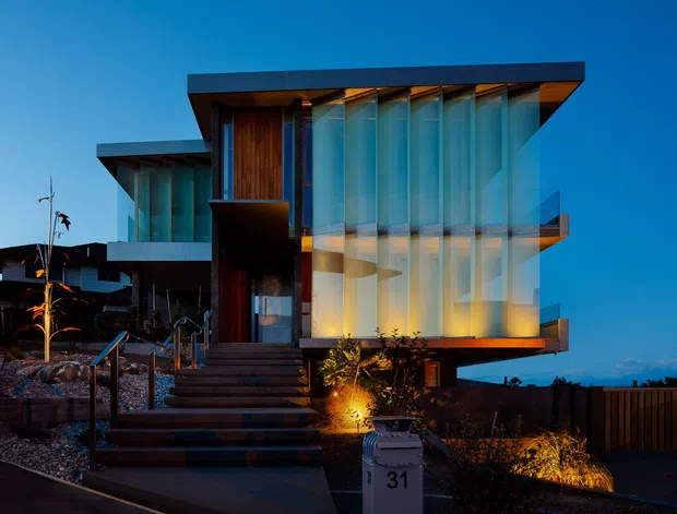 Twilight exterior view of a striking contemporary multi-level residence. The building features a flat, projecting roof, extensive translucent or reflective floor-to-ceiling glazing, dark structural framing, warm timber cladding, and a raised entry approached by broad steps. Landscape lighting highlights the entry planting and facade, while neighbouring residential forms and fencing are visible in the background.