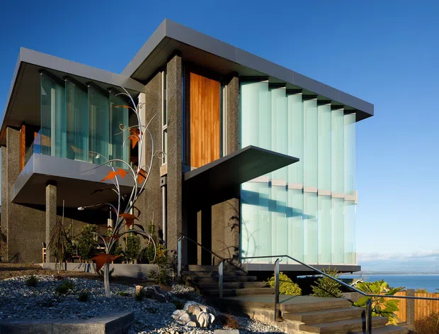 A striking multi-level contemporary coastal residence with strong geometric forms, projecting flat roof planes, dark textured exterior walls, warm timber feature cladding, and extensive floor-to-ceiling glazing with vertical fins. The building sits on a rocky, landscaped sloping site overlooking a body of water, with stepped pathways and terraces leading toward the view.
