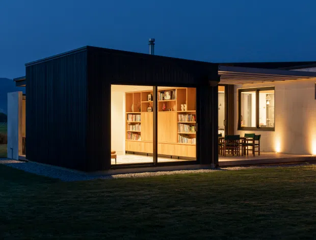 A contemporary single-storey home photographed at dusk, with a dark vertically clad rectangular volume containing a warmly lit library or study behind full-height glazing. An adjoining lighter pavilion opens toward a covered outdoor dining area. The building sits in a rural or semi-rural landscape with distant hills, and the strong contrast between the dark exterior and warm interior lighting creates a dramatic evening atmosphere.