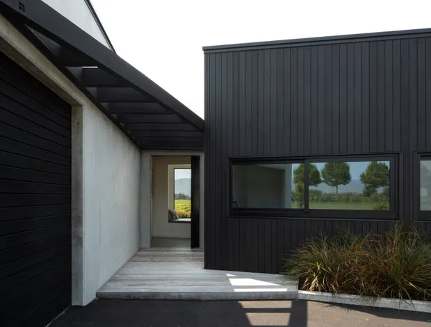 Exterior view of a contemporary single-storey building entrance and covered walkway. The facade combines dark vertical metal cladding, a black sectional garage door, exposed concrete or rendered walls, and horizontal glazing. Native grasses soften the edge of the paved approach, while a landscaped rural outlook is visible through the windows.