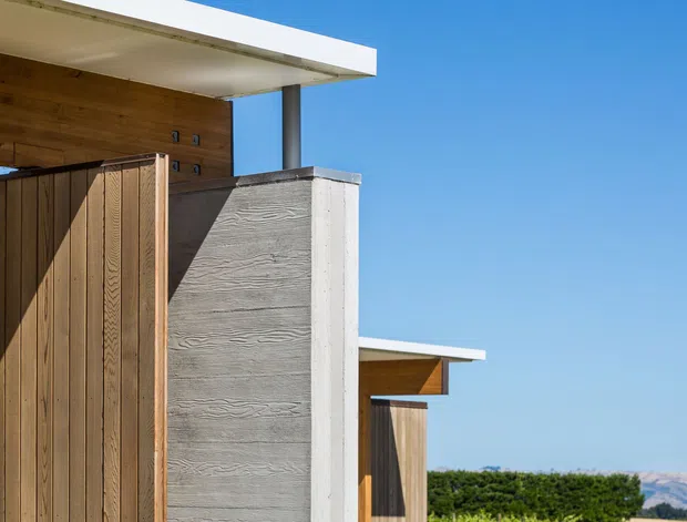 Close-up exterior architectural photograph showing a modern, flat-roofed building with crisp white roof slabs, warm timber cladding and fencing, a grey textured concrete wall, and dark slender structural posts. Clear blue sky and distant landscape are visible in the background.