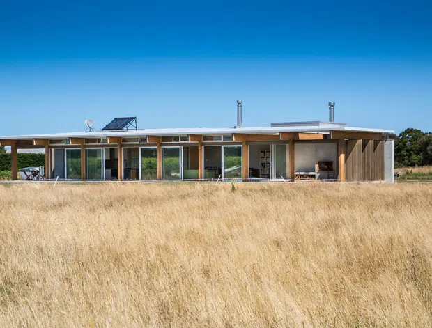 Single-storey contemporary pavilion-style house set within a dry grassy landscape. The elongated building has a very low-pitched or nearly flat roof with broad eaves, extensive floor-to-ceiling glazing, timber-framed openings, and vertical timber-clad sections. A covered outdoor living area is visible at the right, with a restrained linear facade and strong connection to the surrounding field.