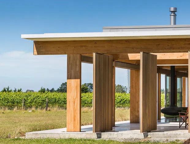 A compact modern pavilion-like building set beside a vineyard, with a flat roof, deep timber soffit, exposed timber posts, and a series of tall vertical timber pivot screens framing full-height glazing. A dark sculptural hanging fireplace and chairs are visible inside. The structure sits on a pale concrete slab and is photographed in bright daylight against a clear blue sky.