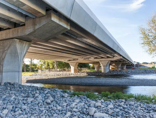 A low-angle exterior view beneath a large concrete bridge spanning a shallow rocky waterway. Repeated sculptural concrete piers support the broad deck, with exposed structural beams and a metallic edge fascia visible along the bridge. The surrounding landscape includes gravel banks, shallow water, trees, and a distant road infrastructure.