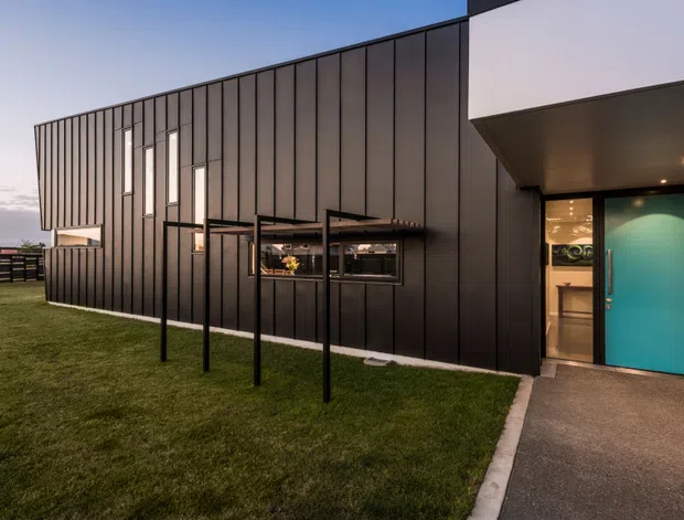 Contemporary single-storey building exterior at dusk, clad in dark vertical metal panels with a projecting entry canopy, turquoise feature door, narrow vertical windows, and a covered outdoor structure along the facade. The building sits beside a neatly landscaped lawn and paved entry path.