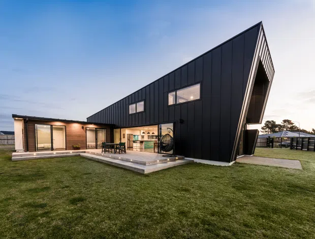 Exterior view of a striking contemporary single-storey home with a dramatic steeply angled gable-like roof form and dark vertical metal cladding. Large glazed openings connect the living areas to a raised concrete patio and broad lawn, with warm interior lighting visible at dusk.
